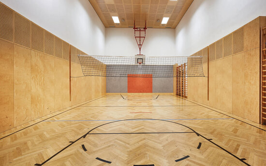 Ein heller Sporthalle mit Holzfußboden, einem Volleyballnetz und einer Basketballanlage an der Wand.