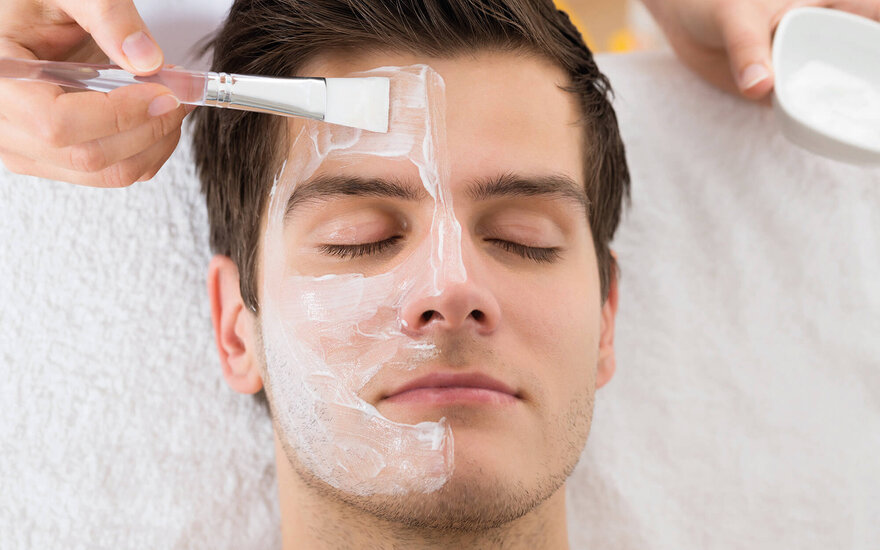 Ein junger Mann erhält eine Gesichtsbehandlung, während eine Hand eine Maske auf sein Gesicht aufträgt.