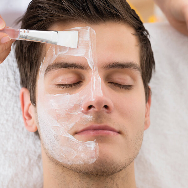 Ein junger Mann erhält eine Gesichtsbehandlung, während eine Hand eine Maske auf sein Gesicht aufträgt.