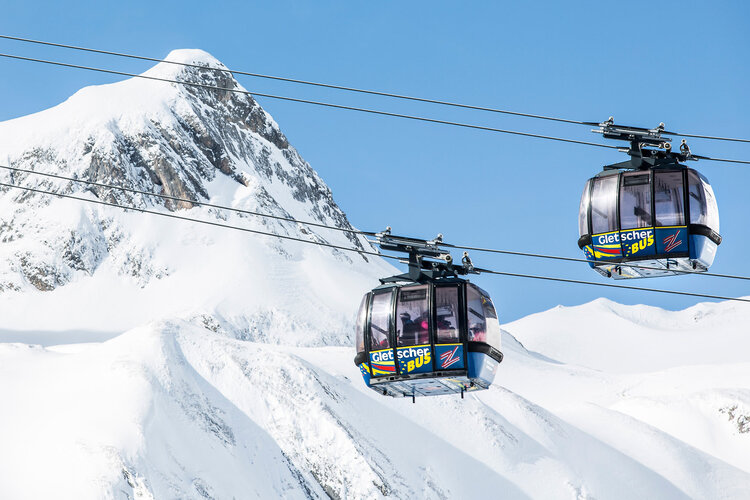 Zwei moderne Gondeln schweben über schneebedeckte Berge, während der klare Himmel im Hintergrund strahlt.