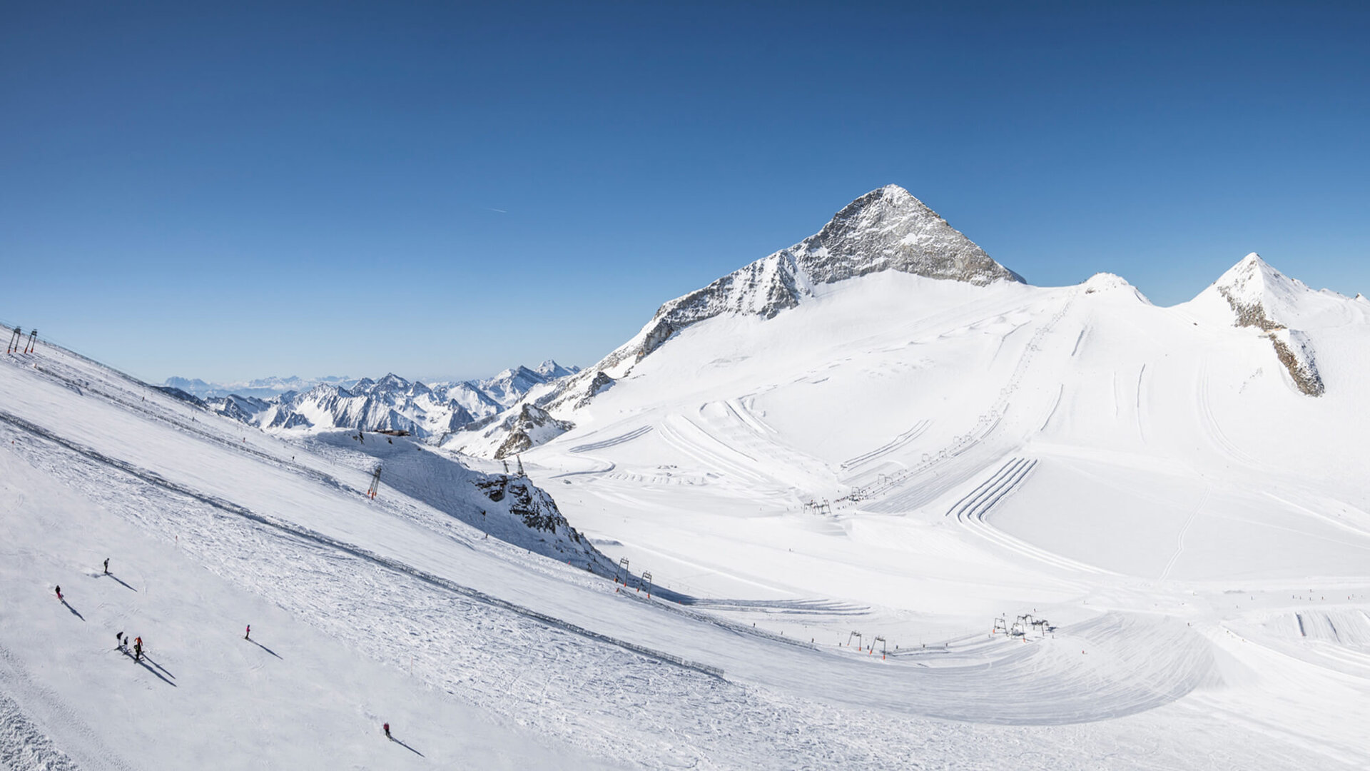 Eine weite, schneebedeckte Berglandschaft mit Skifahrern, die die Pisten hinunterfahren, unter einem strahlend blauen Himmel.
