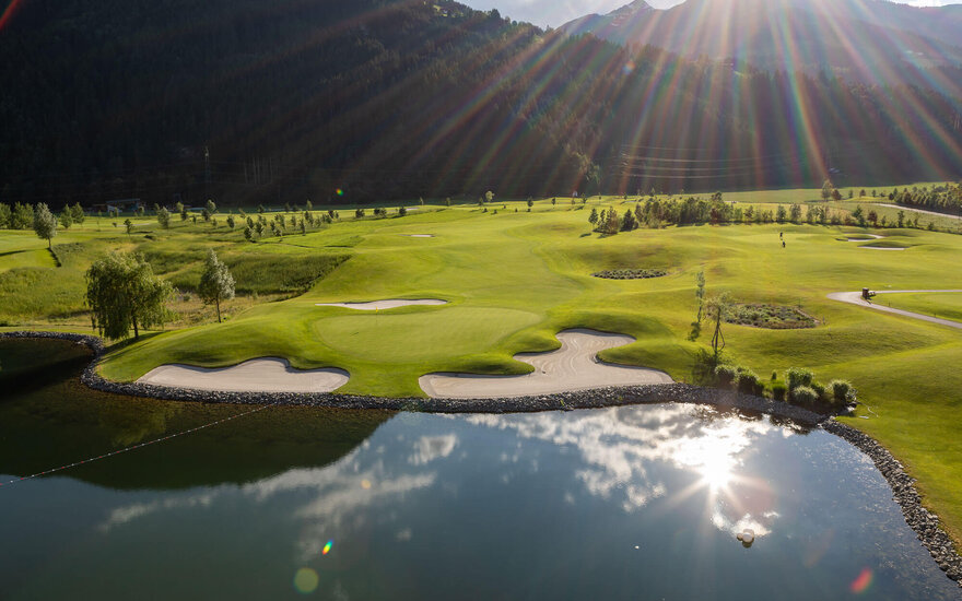 Eine malerische Golfplatzlandschaft mit sanften Hügeln, Sandbunkern und einem ruhigen See, der die Wolken reflektiert.