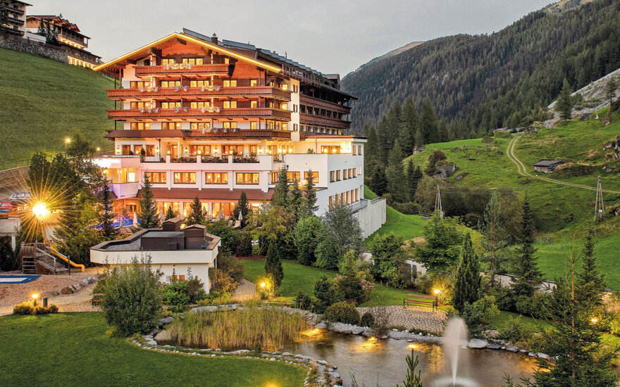 Ein elegantes Hotel mit mehreren Etagen, umgeben von grünen Hügeln und einem kleinen Teich im Vordergrund.