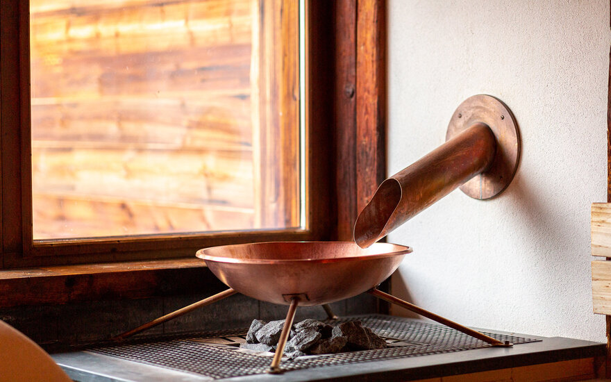 Ein kupferner Aufgussbehälter steht auf einem Steingrill, während Sonnenlicht durch das Fenster strömt.