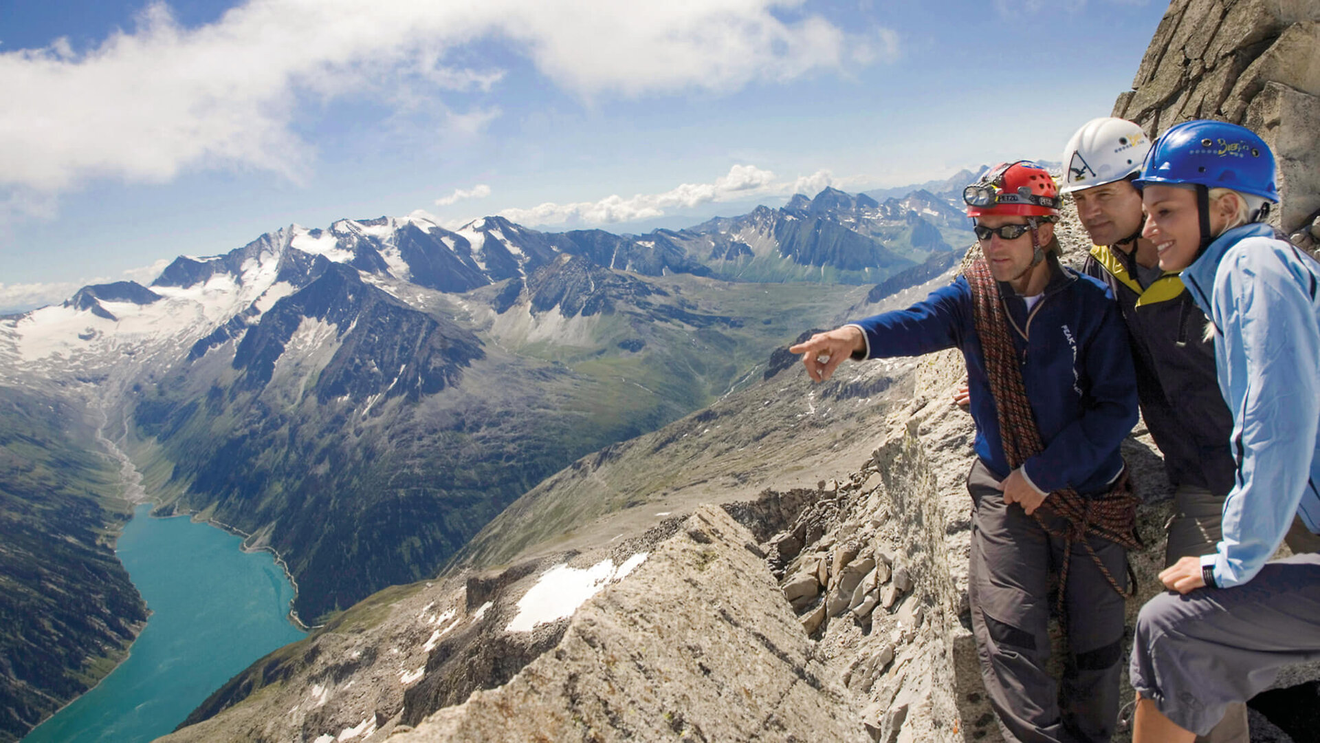 Eine Gruppe von Bergsteigern steht auf einem Gipfel und blickt auf die beeindruckende Berglandschaft und den darunter liegenden See.