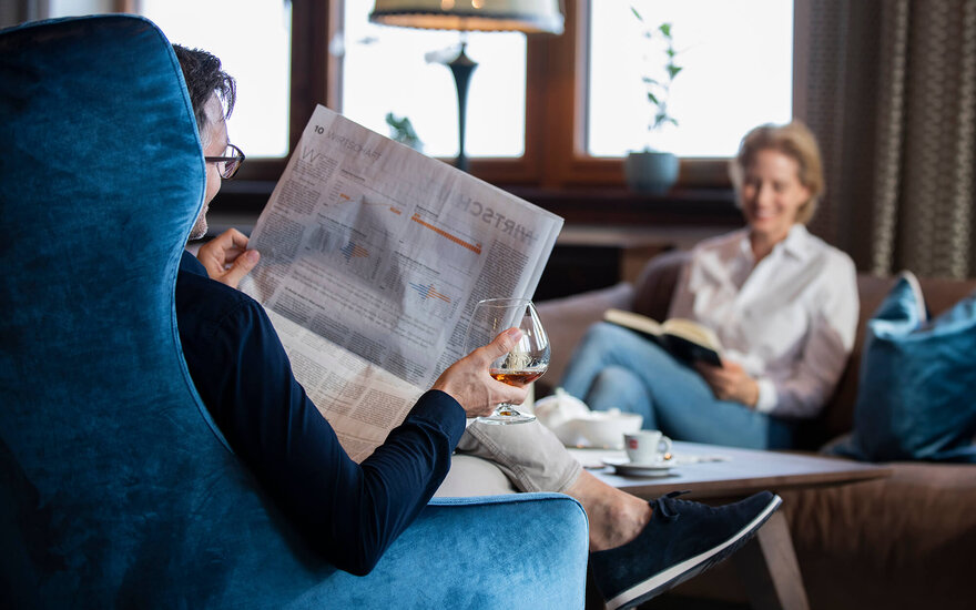 Im Hintergrund einer stilvollen Einrichtung liest eine Frau, während ein Mann in einem Sessel die Zeitung studiert und Whisky genießt.