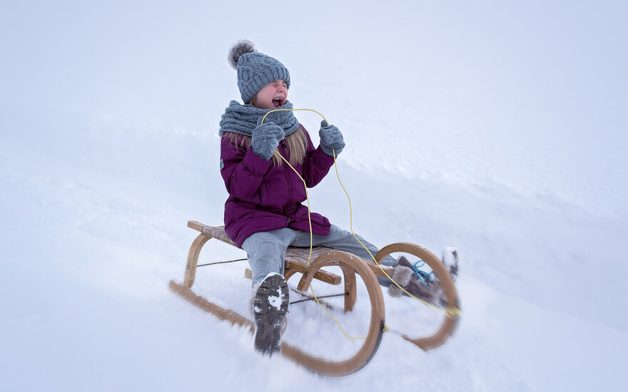 Ein fröhliches Kind in einem lila Wintermantel sitzt auf einem Schlitten und genießt den Schnee mit einem breiten Lächeln.