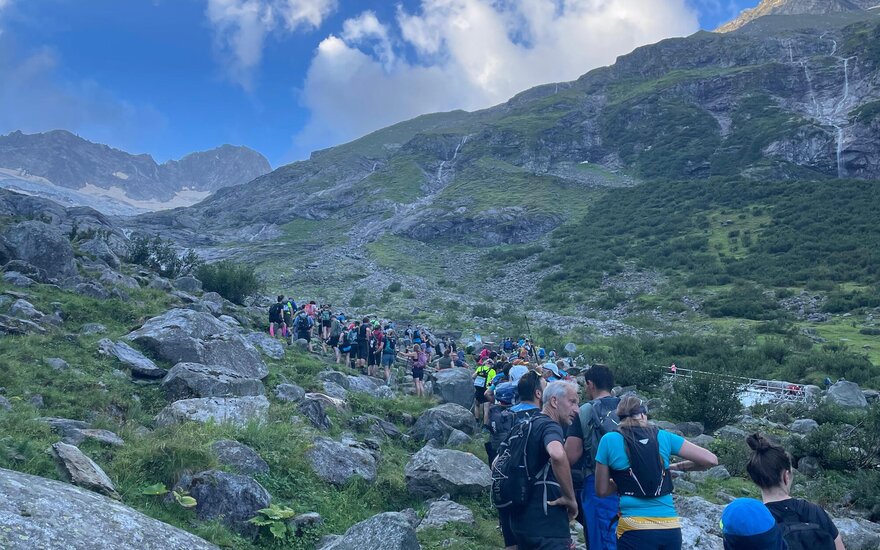 Eine große Gruppe von Wanderern bewegt sich auf einem steinigen Pfad durch eine grüne Berglandschaft unter blauem Himmel.