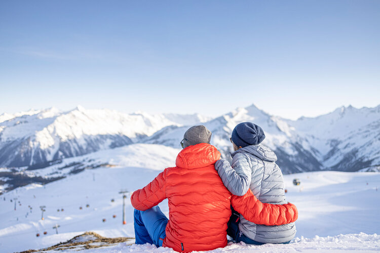 Ein Paar sitzt im Schnee und genießt die atemberaubende Aussicht auf die schneebedeckten Berge im Hintergrund.