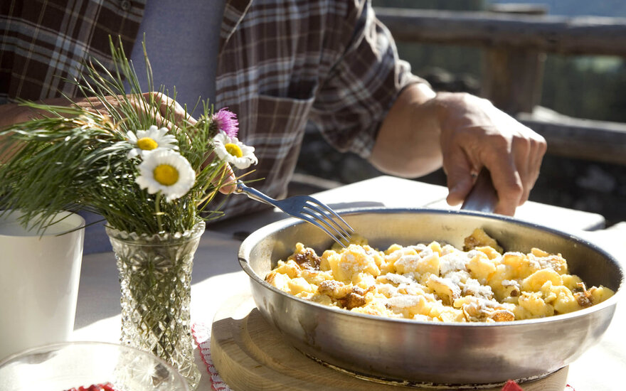 Ein Mann genießt Kaiserschmarrn in einer sonnigen Berghütte, umgeben von frischen Blumen und einer Tasse Kaffee.