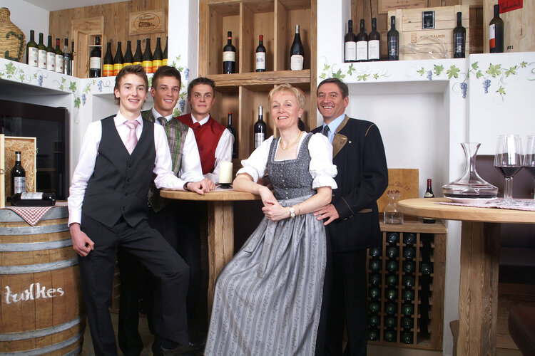 In einem Weinkeller stehen vier Männer und eine Frau, die traditionelle Kleidung tragen, umgeben von Weinregalen und Dekoration.
