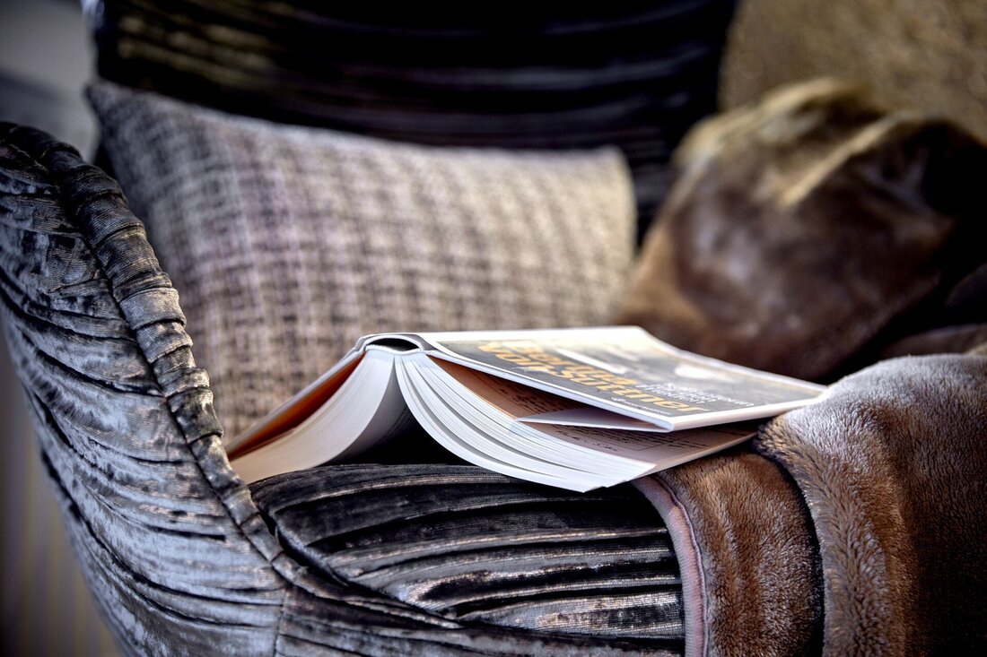 Ein aufgeschlagenes Buch liegt auf einem eleganten, gepolsterten Sessel, umgeben von einem weichen, braunen Überwurf.