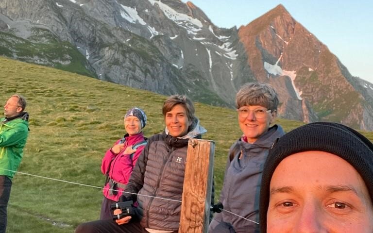 Eine Gruppe von fünf Personen posiert fröhlich vor einer beeindruckenden Berglandschaft bei Sonnenaufgang.