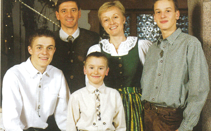 Eine fröhliche Familie posiert in traditioneller Kleidung vor einem rustikalen Hintergrund mit dekorativen Elementen.