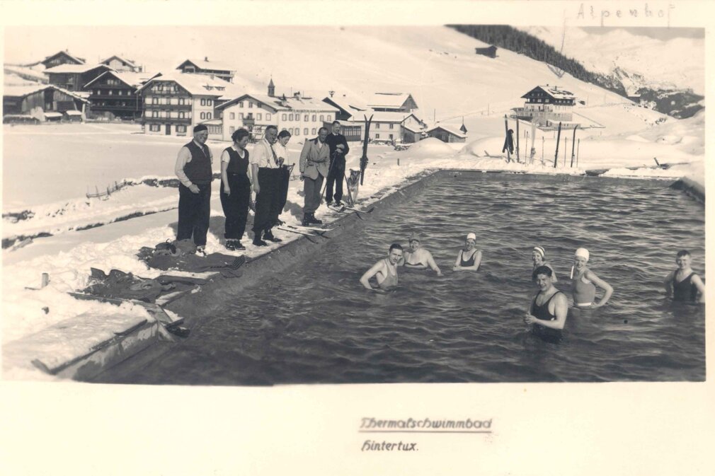 Eine Gruppe von Männern und Frauen genießt das Thermalbad, während die winterliche Landschaft mit schneebedeckten Hügeln im Hintergrund leuchtet.