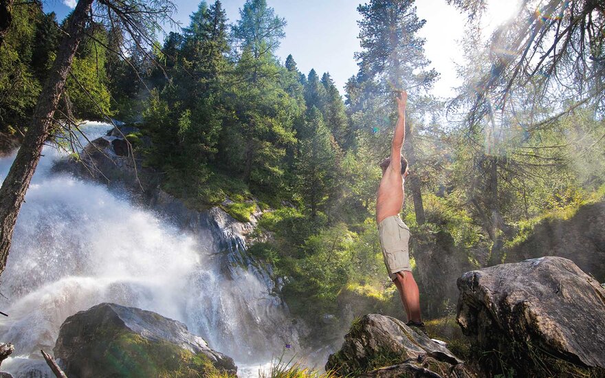 Ein Mann steht auf einem Felsen, hebt die Arme in die Luft und genießt die frische Luft eines Wasserfalls im Wald.