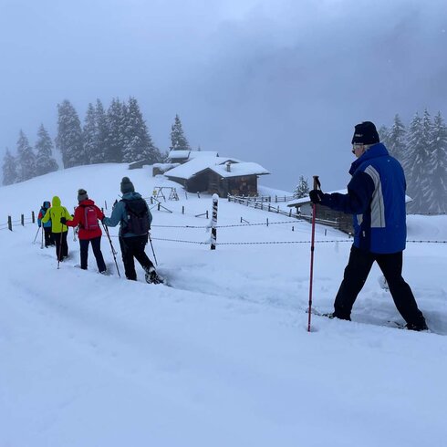 Eine Gruppe von Menschen wandert mit Schneeschuhen durch eine verschneite Landschaft, umgeben von Bäumen und Hütten.