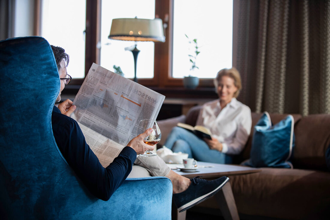 Ein Mann sitzt in einem blauen Sessel, liest eine Zeitung und genießt ein Glas Whisky, während eine Frau im Hintergrund liest.