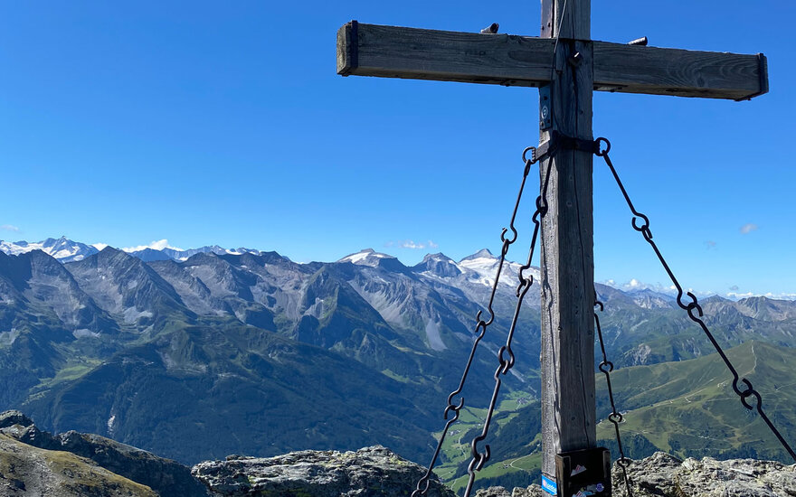 Ein Holzkreuz steht majestätisch auf einem Gipfel, umgeben von beeindruckenden Berglandschaften und klarem blauen Himmel.