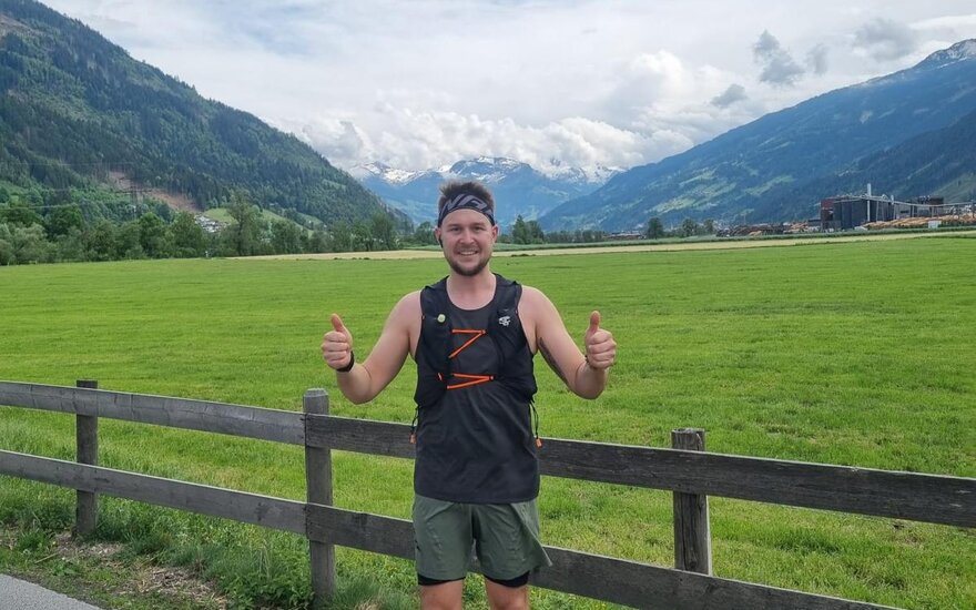Ein junger Mann steht auf einem Weg, lächelt und zeigt Daumen hoch, umgeben von grünen Wiesen und Bergen im Hintergrund.