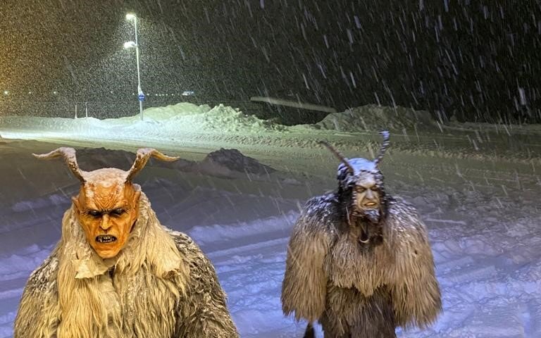 Zwei furchterregende Krampusse in pelzigen Kostümen stehen im Schnee, während Schneeflocken heftig fallen.