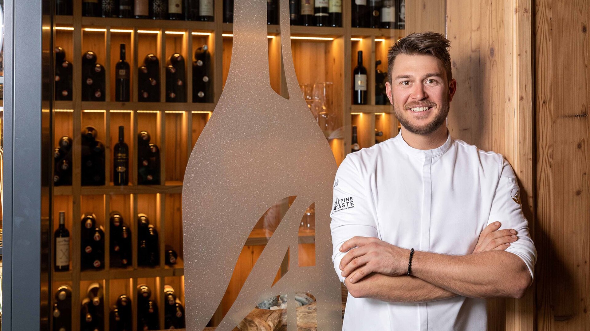 Ein lächelnder Koch in einem weißen Outfit steht vor einer Wand mit beleuchteten Weinflaschen in einem rustikalen Ambiente.