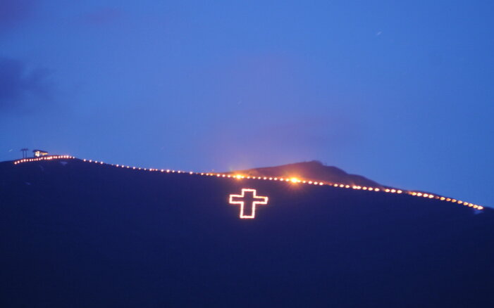Ein beleuchtetes Kreuz auf einem Berggipfel strahlt in der Dämmerung, umgeben von einer Reihe kleiner Lichter.
