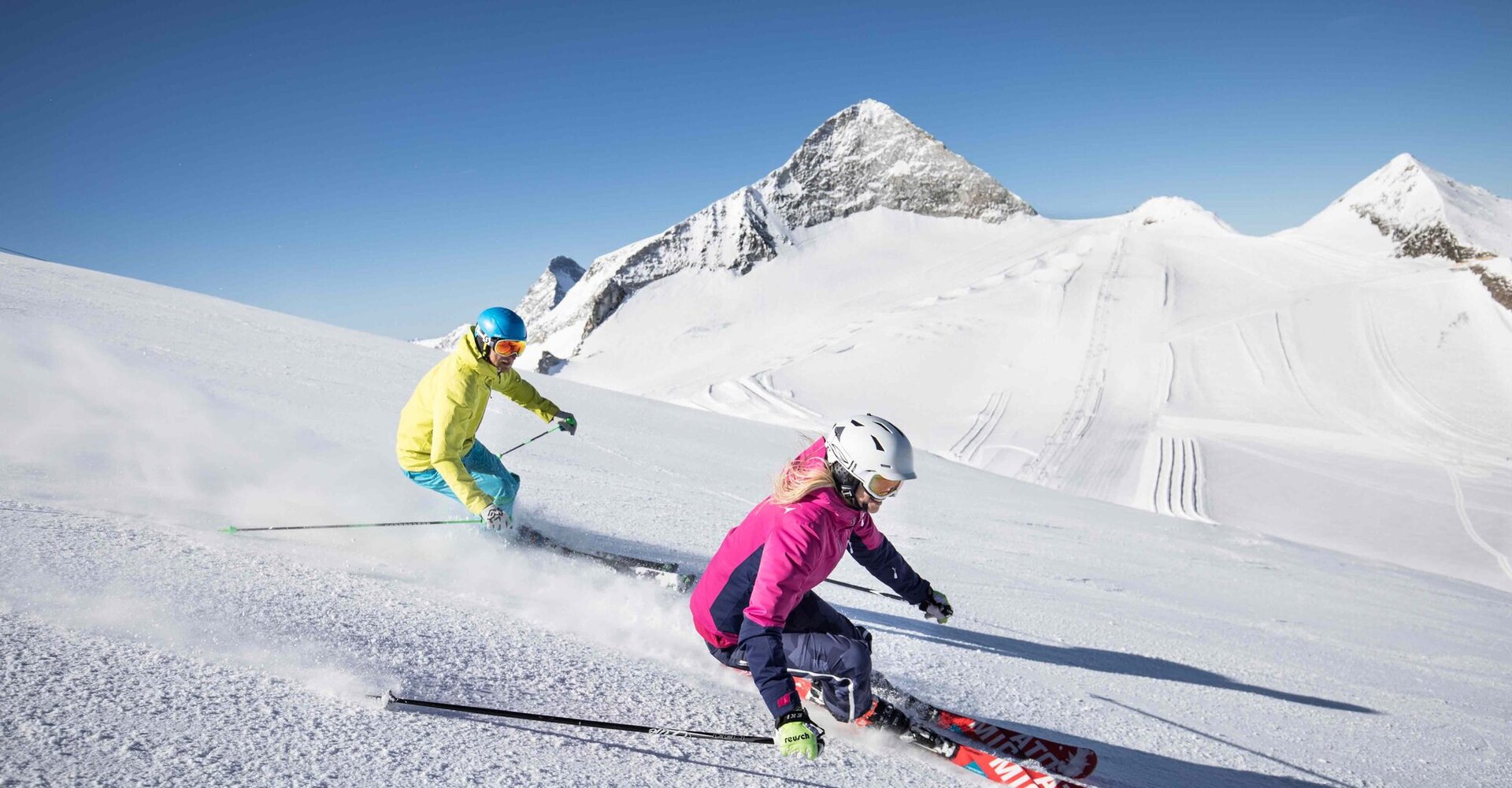 Zwei Skifahrer, ein Mann in gelber Jacke und eine Frau in pinker Jacke, rasen über die schneebedeckte Piste unter klarem Himmel.