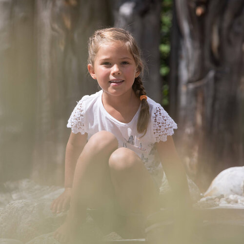 Ein fröhliches Mädchen sitzt auf einem Stein und lächelt, während ein Junge in der Nähe neugierig zuschaut.