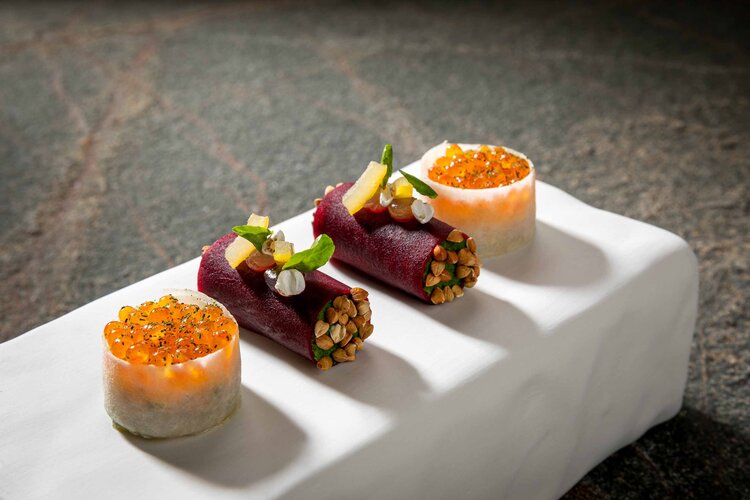 Drei kunstvoll angerichtete Speisen auf einem weißen Teller, darunter Röllchen mit buntem Gemüse und einer Garnitur aus Kräutern.