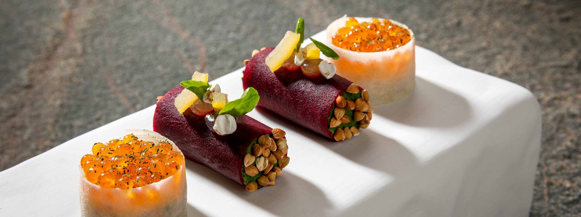 Drei kunstvoll angerichtete Speisen auf einem weißen Teller, darunter Röllchen mit buntem Gemüse und einer Garnitur aus Kräutern.