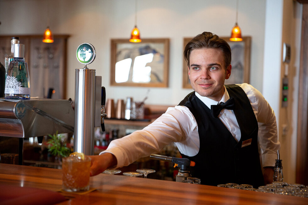 Ein junger Barkeeper in eleganter Kleidung reicht ein Cocktailglas mit einem Lächeln an den Gast weiter.