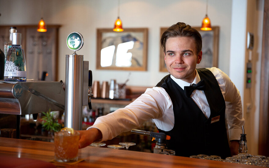 Ein junger Barkeeper in eleganter Kleidung reicht ein Cocktailglas mit einem Lächeln an den Gast weiter.