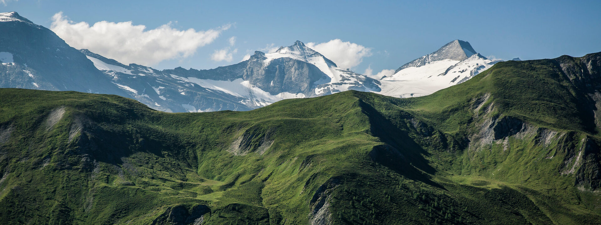 Majestätische Berglandschaft mit schneebedeckten Gipfeln und saftig grünen Hügeln unter einem klaren blauen Himmel.