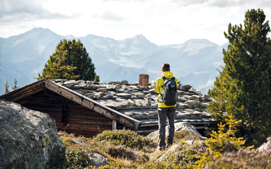 Ein Wanderer in gelber Jacke steht vor einer Berghütte und blickt auf die majestätischen Alpen im Hintergrund.