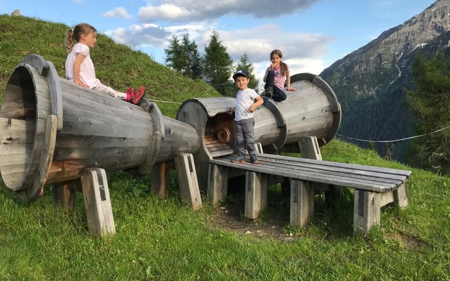 Drei Kinder spielen fröhlich auf einer Holzspielanlage in einer malerischen Berglandschaft mit grünen Wiesen.
